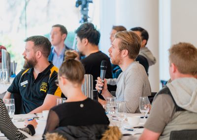 Business workshop attendees seated at tables, with one participant holding a microphone to ask a question