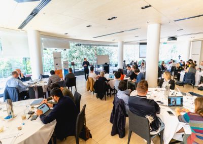 Business workshop with 30+ attendees seated at round tables listening to a presenter at the front of a modern conference room
