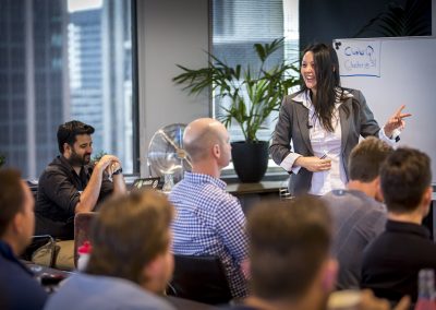Female presenter speaking to a seated group of professionals in a modern boardroom during a business workshop