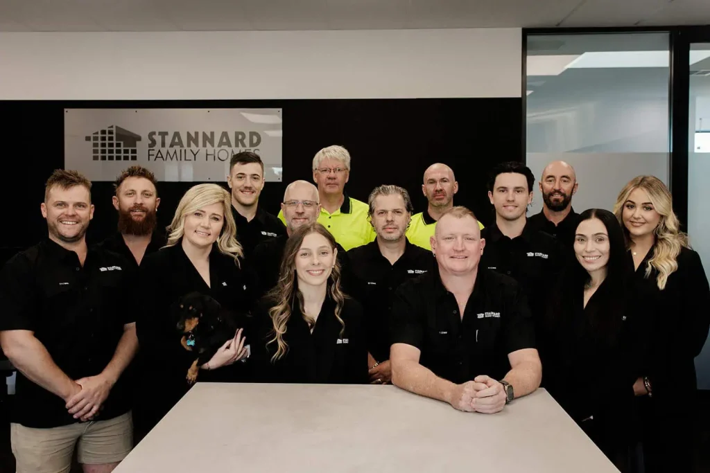 Stannard Family Homes team of 14 staff members posing in their office in front of the company logo sign